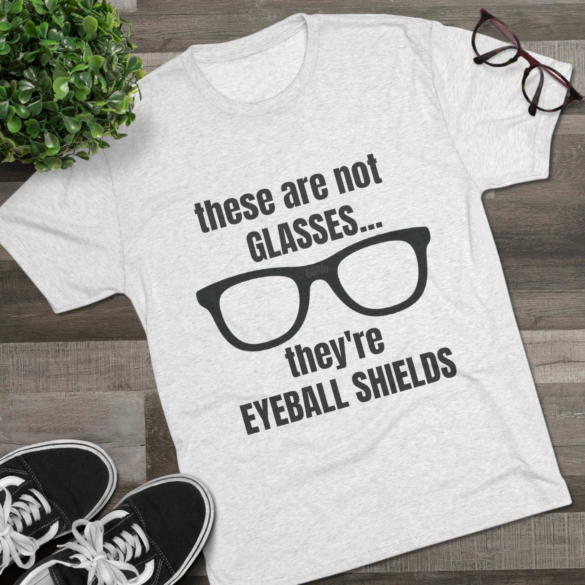 Light heather tri-blend tee flat-laid, printed with black glasses graphic and text ‘these are not GLASSES... they’re EYEBALL SHIELDS.
